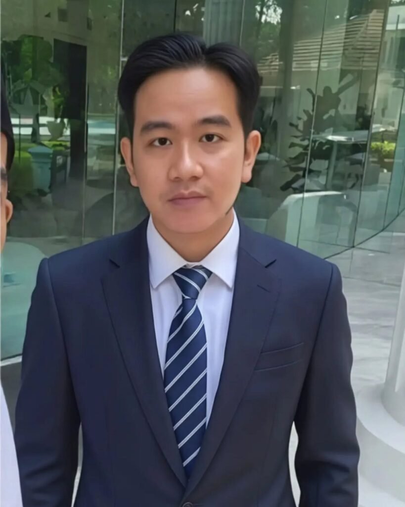 wapres ke 6 gibran mengenakan setelan jas biru tua dan dasi garis-garis, berdiri di depan gedung kaca modern dengan ekspresi wajah serius diterpa isu pemakzulan wapres gibran